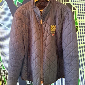 Quiltad jacka från Morris XL - Snygg quiltad jacka från Morris i mörkblått med guldbroderad logga och brittisk flagga på bröstet. Jackan har dragkedja framtill, ståkrage och diskreta fickor. Perfekt för dig som gillar klassisk stil med modern touch. Köpt från Care of Carl. Använt, men bra skick.