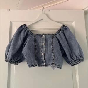 Jeans bolerotopp från GinaTricot  - Superfin croppad bolerotopp i jeans från GinaTricot. Toppen har offshoulder-modell med puffiga korta ärmar och knappar framtill. Perfekt för dig som vill sticka ut med en cool denimlook. Elastiska detaljer vid ärmslut och halsringning.
