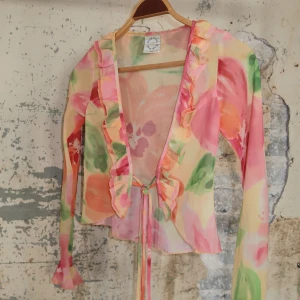 Blommig volangblus med knytband - Superfin blus i transparent material med stora blommor i rosa, orange, grön och gul. Djup v-ringning med volangkant och knytband framtill. Långärmad och lätt croppad modell, perfekt för en färgglad look.