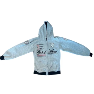 Grå hoodie Geographical Norway L - Grå hoodie från Geographical Norway med broderade detaljer och tryck både fram och bak. Stor siffra 5 på ryggen, texten Cote d’Azur och Monte Carlo, samt patchar på ärmarna. Svarta muddar och huva med dragsko. Mjuk insida i bomullsblandning. Passar S-M. Har färgfläck på ena armmudden