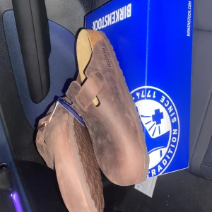 Bruna Birkenstock Boston i mocka - Säljer ett par Birkenstock Boston i brun mocka med justerbart spänne i metall och korksula. Skorna har en klassisk slip-in design och är perfekta för dig som gillar bekväma och stilrena sandaler. Märket är präglat på sidan och sulan är mönstrad för bra grepp. Storlek 42 oanvända från Zalando 