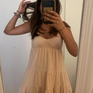 Ljusbeige sommarklänning med volanger - Supersöt ljusbeige klänning med tunna axelband och v-ringning. Klänningen har flera volanglager och en luftig passform, perfekt för varma dagar. Nederdelen har en fin spetsdetalj som ger extra sommarkänsla. Materialet känns lätt och skönt mot huden.