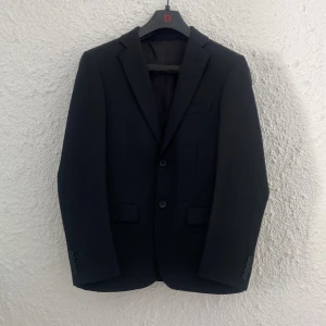 Svart kavaj med röd detalj från Dressmann - Stilren svart kavaj med klassisk passform och två knappar framtill.  Används endast på ett par bröllop och 9:ans bal 