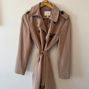 Beige trenchcoat från H&M, stl 34 - Snygg beige trenchcoat från H&M i klassisk modell med knytskärp i midjan och svarta knappar. Jackan har axelklaffar, krage och långa ärmar. Perfekt för vår och höst, passar till allt och ger en clean look.