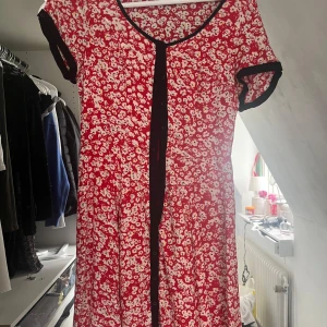 Röd blommig klänning River Island - Supersöt röd kortklänning från River Island med vita blommor och svarta detaljer vid ärmslut och knäppning. Klänningen har korta ärmar, rund halsringning och knappar framtill. Perfekt för dig som gillar färg och mönster!