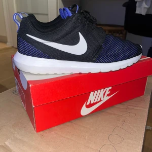 Svarta och blå Nike sneakers - Snygga Nike sneakers med svart mesh-överdel och blått mönster, vit sula och klassisk vit swoosh på sidan. Skorna har snörning och en sportig, modern look som passar perfekt till streetwear. Lätta och bekväma med rund tå och platt sula.