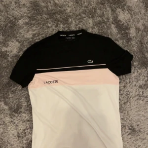 Lacoste t-shirt i svart, rosa och vitt - Snygg t-shirt från Lacoste med svart överdel, ljusrosa mittparti och vit nederdel. Klassisk krokodil-logga på bröstet och Lacoste-tryck. T-shirten har korta ärmar och är gjord i mjuk bomull, perfekt för en clean och sportig look.