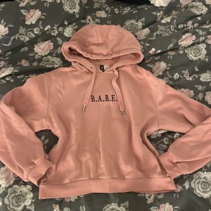 Rosa hoodie  - Säljer en ljusrosa hoodie från Divided i storlek XS med svart texten 'B.A.B.E.' framtill. Tröjan har huva med snörning och långa ärmar. Perfekt för dig som gillar en chill och snygg stil. Materialet är mjuk bomullsmix. 