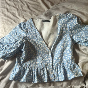 Blommig blå blus från Zara - Supersöt blommig blus från Zara i ljusblått med vita små blommor. Modellen har v-ringning, knappar framtill, volangkant nertill och puffiga trekvartsärmar med resår. Perfekt för vår och sommar när du vill ha en romantisk vibe.