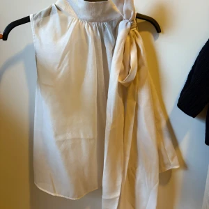 Vit ärmlös blus med knyt - Stilren vit blus i glansigt material med hög hals och snygg knytning vid axeln. Den är ärmlös och har ett lätt, luftigt fall som ger en elegant look. Perfekt för dig som gillar en chic och modern stil.
