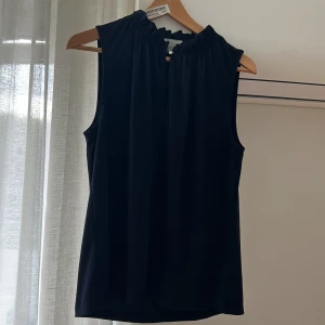 Svart ärmlös blus med rynkad krage från HM - Stilren svart blus med rynkad hög krage och ärmlös design. Perfekt att matcha med jeans eller kjol för en clean look. Lätt och luftig känsla, passar dig som gillar minimalistisk stil.