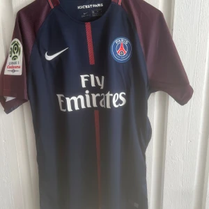 PSG Neymar Jr matchtröja blå/röd - Paris Saint-Germain fotbollströja med Neymar Jr och nummer 10 på ryggen. Tröjan är marinblå med vinröda ärmar, vita tryck och röda detaljer. Officiell Nike Dri-Fit med Fly Emirates sponsorlogga och Ligue 1-märke på ärmen. Perfekt för fans av PSG och Neymar.