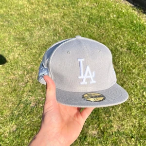 Grå LA Dodgers keps New Era  - Snygg grå LA Dodgers keps från New Era 59FIFTY med broderad LA-logga framtill och 1988 World Series-detalj på sidan. Klassisk flat peak och broderade ventilationshål. Perfekt för dig som gillar streetwear och sportig stil. Köparen betalar frakt