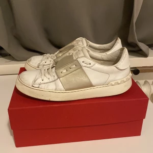 Valentino Garavani Open sneakers vit/silver - Snygga Valentino Garavani Open sneakers i vitt skinn med guld detalj över mitten och ikoniska nitar baktill. Klassisk rund tå, platt sula och snörning framtill. Perfekta för dig som vill ha en lyxig och trendig look passar inte priset kom med prisförslag