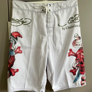  vita shorts med dödskalle - Snygga vita shorts med stora, färgstarka dödskalle- och ormtryck i rött, blått och svart. Shortsen har snörning i midjan och en bakficka med lock. Perfekta för dig som vill sticka ut med edgy stil. Midjemått 80 cm 