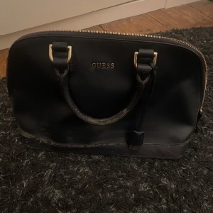 Svart handväska från Guess - Stilren svart handväska från Guess med guldfärgade detaljer och dragkedja upptill. Priset går att diskutera pågrund av slitningarna så ge gärna prisförslag!! (Jag ska rengöra den den har stått och dammat lite men har inte hunnit med!)