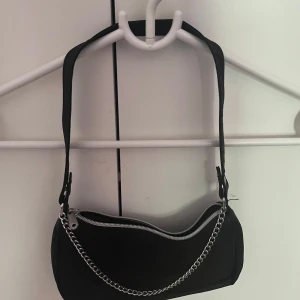 Svart axelväska med kedjedetalj - Säljer en svart axelväska i syntetmaterial med silvrig dragkedja och en snygg kedjedetalj framtill. Väskan har en enkel, stilren design och en justerbar axelrem. Perfekt storlek för det viktigaste och passar till många olika outfits.