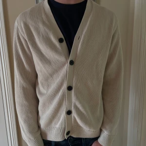 Beige kofta från H&M med knappar - Säljer en stilren beige kofta från H&M i storlek S. Koftan är stickad i bomull, har långa ärmar och stängs med svarta knappar framtill. Perfekt för lager-på-lager och enkel att matcha med jeans eller t-shirt.