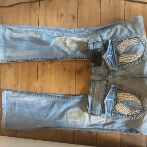 Miss Me ljusblå bootcut jeans strl 27 - Ljusblå jeans från Miss Me med bootcut passform och snygga slitningar på benen. Bakfickorna har coola vingdetaljer med glittriga stenar. Jeansen har dubbla knappar och dekorativa nitar. Perfekta för dig som gillar statement-jeans med extra detaljer.