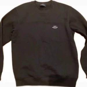 Svart sweatshirt från Mads Nørgaard - En stilren svart sweatshirt från Mads Nørgaard med liten vit logga på bröstet. Klassisk rund halsringning och långa ärmar. Tillverkad i mjuk bomull, perfekt för en clean och avslappnad look.