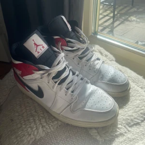 Nike Air Jordan 1 Mid Vit/Röd/Svart - Säljer ett par Nike Air Jordan 1 Mid i klassisk vit, röd och svart färg. Skorna har svart swoosh, röd häl och vit ovandel i skinn. Perforerad tå, högt skaft och ikonisk Jumpman-logga på plösen. Perfekta för dig som gillar streetwear och sneakerskultur.