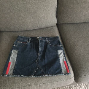 Jeanskjol från Tommy Jeans med detaljer - Mörkblå jeanskjol från Tommy Jeans med rå kant nertill, röda och grå detaljer på sidorna och klassiska fickor både fram och bak. Kjolen har en snygg denimlook och passar perfekt till sneakers eller boots.