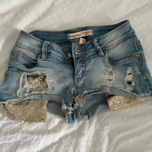 Blå jeansshorts med paljettdetaljer - Snygga ljusblå jeansshorts från Monday Denim med slitna detaljer och råa kanter. Shortsen har guldiga paljetter som sticker fram.. Perfekta för dig som vill sticka ut i sommar!