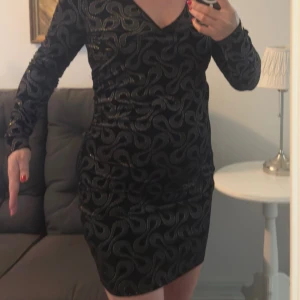 Svart sammetsklänning Chiara Forini - Supersnygg svart sammetsklänning från Chiara Forini Milano med glittrigt mönster i silver och guld. Klänningen har lång ärm, v-ringning och är kort i modellen. Perfekt för dig som vill sticka ut med en glamorös look.