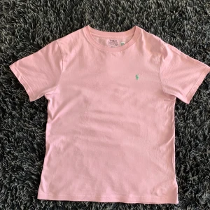 Rosa t-shirt från Polo Ralph Lauren - Snygg rosa t-shirt från Polo Ralph Lauren med klassisk passform och ärmar. T-shirten har en broderad grön logga på bröstet och är gjord i mjuk bomull. Perfekt för dig som gillar stilrena plagg. Passformen är bra då det passar även för folk mellan 165-170.
