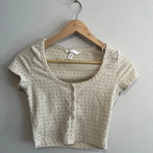 Beige croppad topp med knappar H&M - Superfin beige, kort topp med kofta ärmar och hålmönster över hela tyget. Toppen har en rund halsringning och knappar framtill