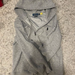 Grå hoodie från Polo Ralph Lauren - Snygg grå hoodie från Polo Ralph Lauren i custom fit. Tröjan har dragkedja, huva med snörning och den klassiska broderade loggan på bröstet. Perfekt för en chill och stilren look. Tillverkad i mjuk bomullsmix för extra komfort. Pris kan diskuteras vid snabb affär.