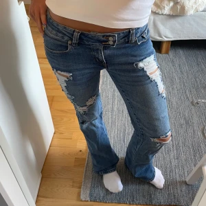 Blå slitna bootcut jeans  - Säljer ett par blå jeans med bootcut passform och slitna detaljer på låren och knäna. Jeansen har låg midja och klassisk femficksdesign. Perfekta för dig som gillar en avslappnad och trendig look.