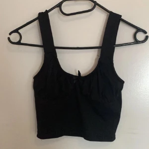 Svart croppad linnetopp H&M Divided XS - Svart croppad linnetopp från H&M Divided med breda axelband och liten rosett framtill. Toppen har en enkel och stilren design med lätt rynkade detaljer vid bysten. Perfekt att matcha med höga jeans eller kjol.