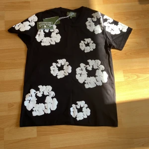 Svart t-shirt med vita blommor Denim Tears - Svart t-shirt från Denim Tears med stora vita blommiga tryck över hela plagget. Klassisk passform med rund hals och korta ärmar. Tillverkad i mjuk bomull som känns skön mot huden. Perfekt statement-piece för dig som vill sticka ut.