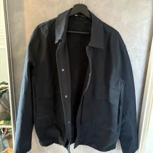 Svart overshirt från Zara - Stilren svart overshirt från Zara i bomull med klassisk krage och stora fickor framtill. Jackan har en rak passform och stängs med knappar. Perfekt för lager-på-lager och enkel att matcha med olika outfits.