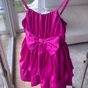 Rosa kort klänning med rosett - Kort klänning i  rosa med justerbara axelband och stor rosett framtill. Klänningen har volangig kjol och är gjord i ett glansigt, satinliknande material som ger en lyxig känsla. 