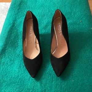 Svarta pumps från Caprice med klack - Stilrena svarta pumps från Caprice med spetsig tå och elegant hög klack. Skorna är i mocka och har en klassisk, tidlös design som passar till många olika outfits. Perfekta för dig som vill ha en chic och sofistikerad look.