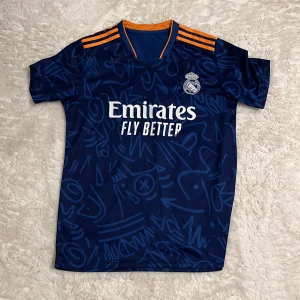 Real Madrid Vini Jr. blå bortatröja - Säljer en blå Real Madrid bortatröja från Adidas med Vini Jr. och nummer 20 på ryggen. Tröjan har orange detaljer på axlarna, korta ärmar och ett coolt grafiskt mönster. Materialet är 100% polyester och trycket på bröstet är Emirates Fly Better.