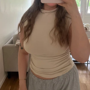 Beige ärmlös polotopp - Säljer en stilren beige polotopp med ärmlös design och tight passform. Toppen har en hög hals och är tillverkad i ett mjukt, stretchigt material som sitter snyggt mot kroppen. Perfekt att matcha med både jeans och kjol för en clean look. Soft touch kollektionen från Gina. 