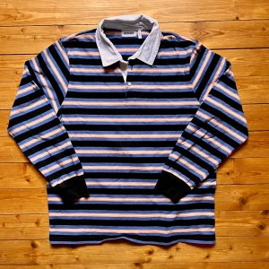 Randig pikétröja från Weekday - Säljer en randig pikétröja från Weekday med klassisk vit krage och knäppning framtill. Tröjan har svarta, blåa och orange ränder, långa ärmar med svarta muddar och är gjord i mjuk bomull. Perfekt för dig som gillar retrovibbar och streetstyle.