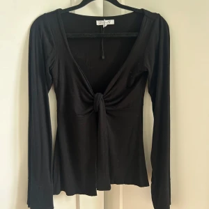 Svart knytblus - Design by Si - Svart långärmad topp från Design by Si med djup v-ringning och knyt framtill. Otroligt mjukt material. Använd kanske 1-2 gånger och säljes pga jobbar numera som lärare.. Ord. Pris: 450 kr.