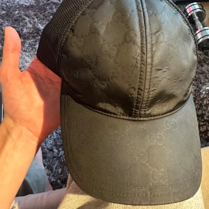 Svart keps från Gucci nylon med mesh storlek M - Snygg svart keps från Gucci med diskret GG-monogram över hela framsidan. Kepsen har mesh på sidorna och bak, samt justerbar snapback. Framsidan är i blankt tyg och baksidan har en läderpatch med Gucci-logga. Perfekt för dig som gillar exklusiva accessoarer.