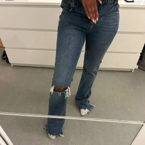 Blå bootcut jeans med slitningar - Säljer ett par blå bootcut jeans med hög midja och slitna detaljer vid knäet och bensluten. Jeansen har klassisk fyraficksdesign och är tillverkade i kraftigt denimtyg. Perfekta för dig som gillar en avslappnad och trendig look.
