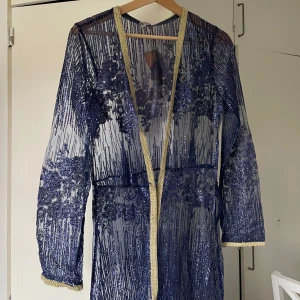 Lång blå glittrig klänning - Säljer en lång blå abaya med transparent mesh och glittriga paljettdetaljer. Man kan ha en klänning under, är en sorts komplement till en klänning, har guldiga kanter längs öppningen och ärmsluten. Är inte en hög kvalite vara. Ej stretchigt men väldigt glittrigt 