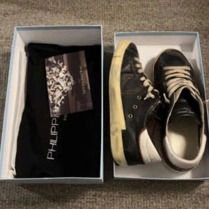 Philippe Model svarta sneakers i skinn - Svarta sneakers från Philippe Model med vit sula och vita snören. Skorna har detaljer i vitt och beige baktill samt diskret logga på sidan. Tillverkade i skinn med klassisk låg profil och rund tå. Perfekta för en stilren och avslappnad look.