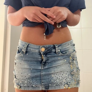 Lågmidjade jeanskjol med blommor - Snygga lågmidjade jeanskjol i ljusblå denim från she wants. Kjolen har blommiga broderier och strassdetaljer på fickorna, samt ett coolt märke i skinn och metall bak. Perfekta för dig som vill sticka ut med detaljer och bling.