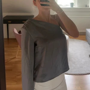 Grå oneshoulder topp i satin - Säljer en stilren grå oneshoulder topp i glansigt satinmaterial. Toppen har en lång ärm på ena sidan och är axelbandslös på den andra. Knappt använd. 