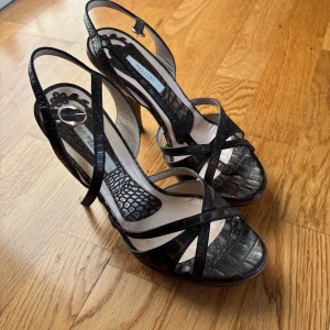 Bruna sandaler med klack från Pollini - Snygga bruna sandaler från Pollini med krokodilpräglat skinn, smala remmar och öppen tå. Skorna har en elegant hög klack i trälook och spänne vid ankeln. Perfekta för dig som vill sticka ut med en stilren och lyxig vibe.