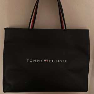 Svart handväska Tommy Hilfiger - Stilren svart handväska från Tommy Hilfiger med klassisk logga framtill. Väskan har rejäla handtag med röd och vit detalj på insidan. Rymmer mycket och är perfekt för dig som vill ha en snygg och praktisk väska till vardagen. köpt för längesedan och därför finns den inte kvar nypris:1199. Använd några gånger men inga tecken på användning eller slitage. 
