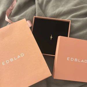 Oandvänt kors Guldarmband från Edblad - Snyggt och stilrent armband från Edblad i guldtonad metall med ett litet kors som detalj. Tunn kedja med justerbar längd och diskret Edblad-logga vid låset. Perfekt för dig som gillar minimalistiska smycken med en twist.